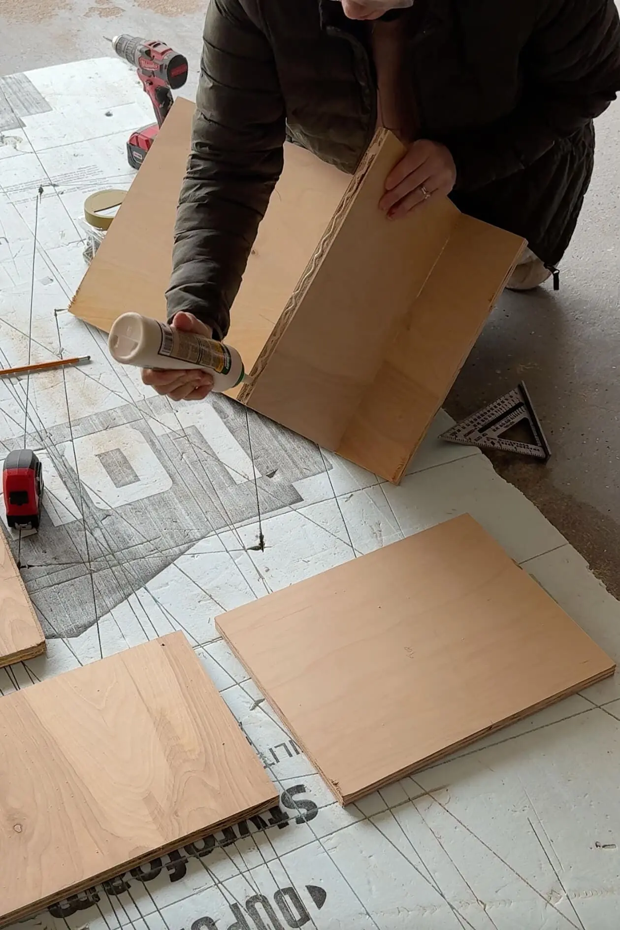 A DIYer in a green puffer jacket applying wood glue to the edge of a plywood board before assembling bedroom built ins. The project pieces are laid out on a foam surface for easy measuring and alignment, with a tape measure, drill, and square nearby.